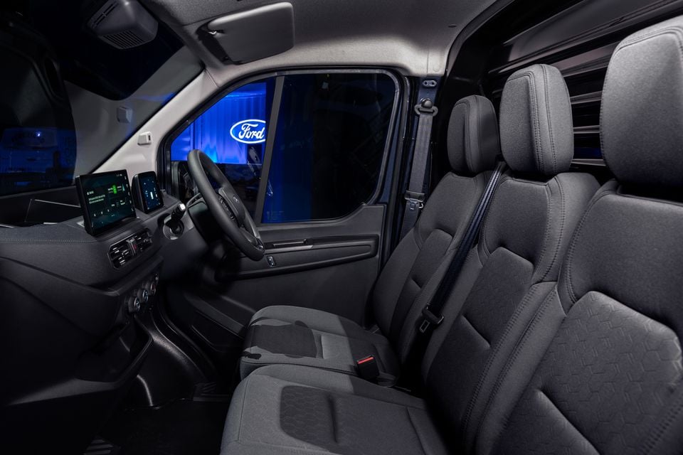 Interior of a Ford Transit City van showing the front cabin with dark fabric seats, steering wheel, and touchscreen infotainment display, with a Ford logo visible through the window