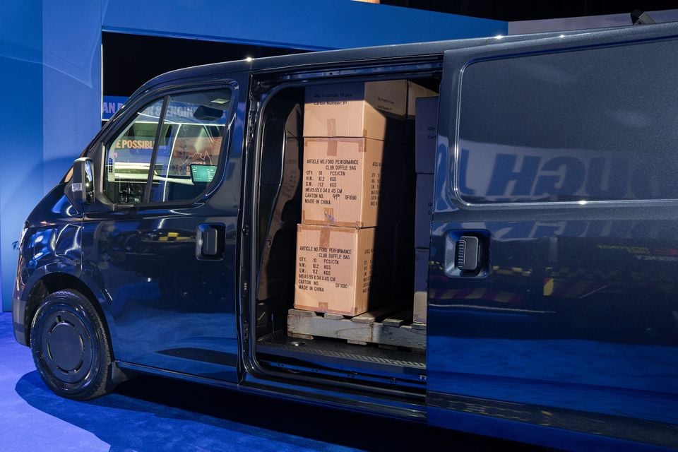 A dark blue 2026 Ford Transit City van with its sliding side door open, revealing cardboard boxes loaded in the cargo area, displayed at a motor show