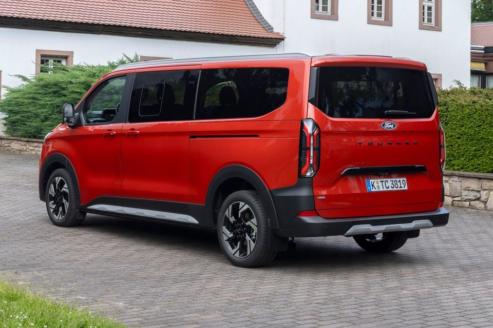 A red Ford Tourneo Custom van seen from the rear three-quarter angle, parked on a brick driveway beside a European building