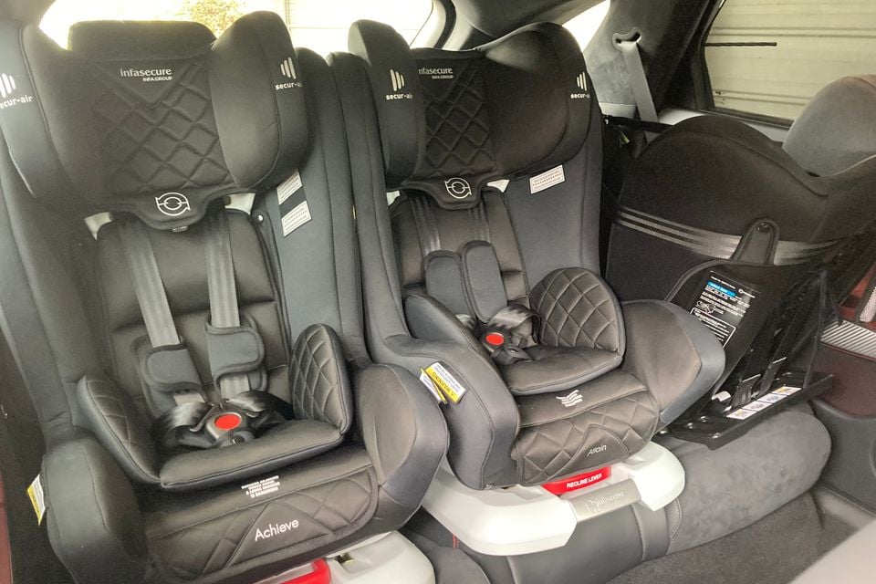 Three child car seats — two Infasecure Achieve and one Infasecure Attain — installed across the rear seat of a car, with a fourth seat visible on the right