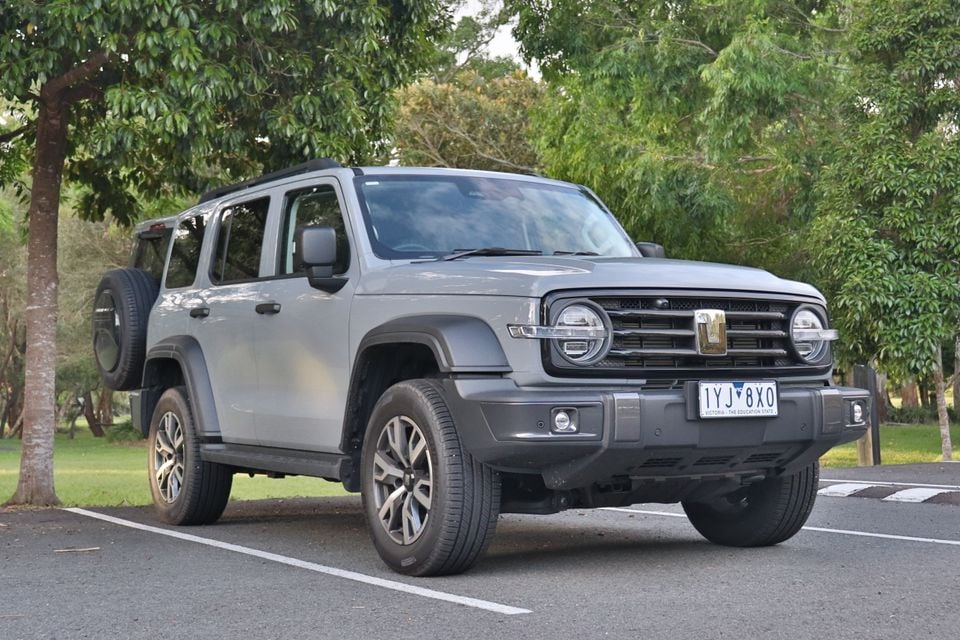 Um SUV cinza 2026 GWM Tank 300 PHEV com roda sobressalente montada na traseira, estacionado em um estacionamento cercado por árvores.