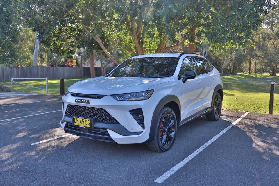 A white 2026 Haval H6GT coupe SUV parked in a car park beside a grassy park, seen from the front three-quarter angle