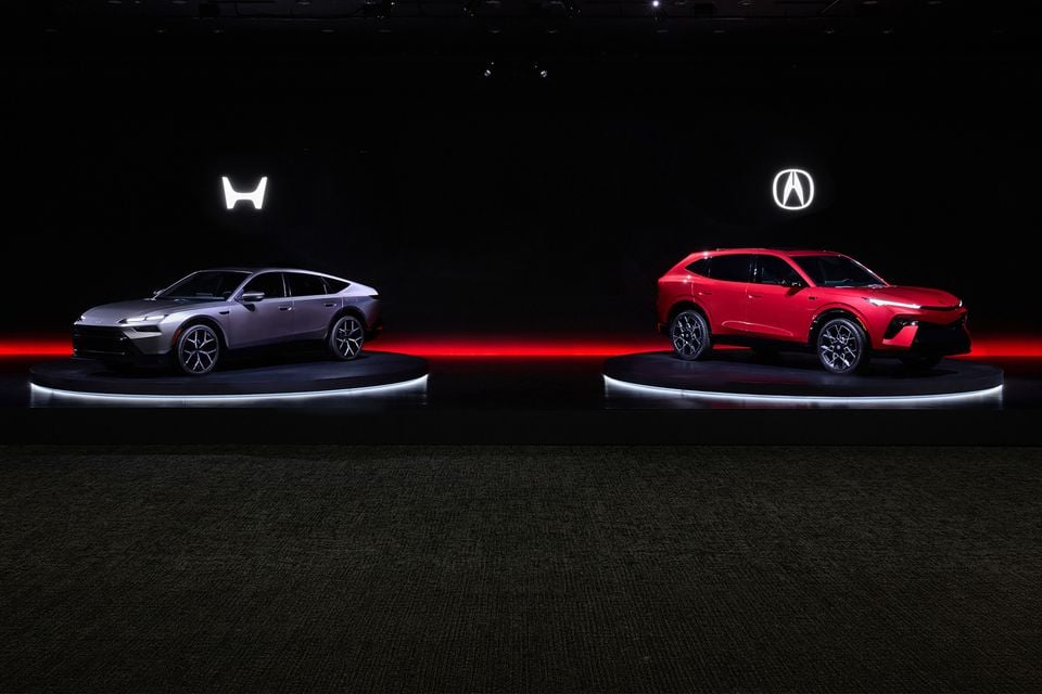 A silver Honda hybrid sedan concept and a red Acura SUV concept displayed on illuminated rotating platforms at a motor show event, with glowing Honda and Acura logos in the background.