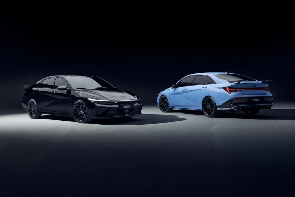 A black 2026 Hyundai i30 Sedan N Monochrome seen from the front beside a blue Hyundai i30 Sedan N seen from the rear in a dark studio