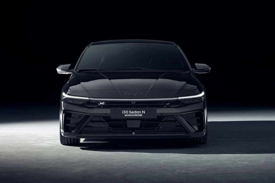 A black 2026 Hyundai i30 Sedan N Monochrome seen from the front in a dark studio, with its full-width light bar and N badge on the grille.