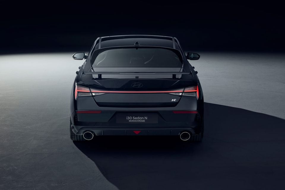 A black 2026 Hyundai i30 Sedan N Monochrome seen from the rear, showing its wing spoiler, full-width tail-light and twin exhausts in a dark studio