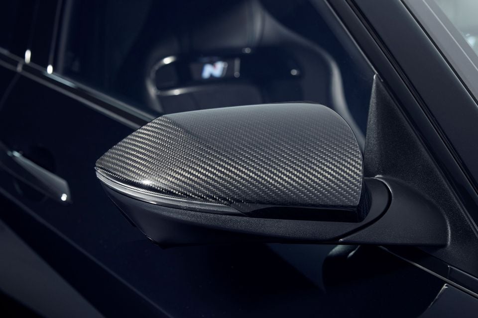 Close-up of the carbon-fibre side mirror cap on a black 2026 Hyundai i30 Sedan N Monochrome, with an N-badged sports seat visible through the window