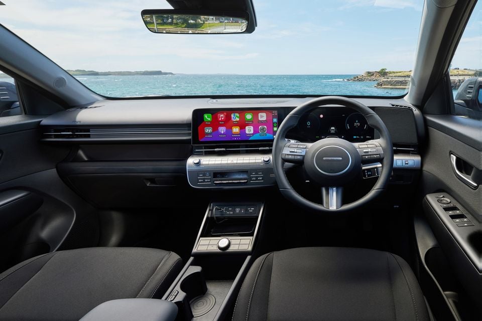 The dashboard and steering wheel of a 2026 Hyundai Kona Electric, with a coastal ocean view through the windscreen.