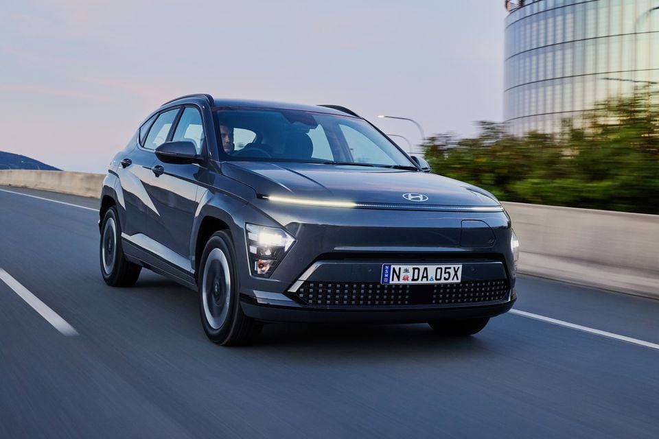 A dark grey 2026 Hyundai Kona Electric SUV driving on a highway, seen from the front quarter view.