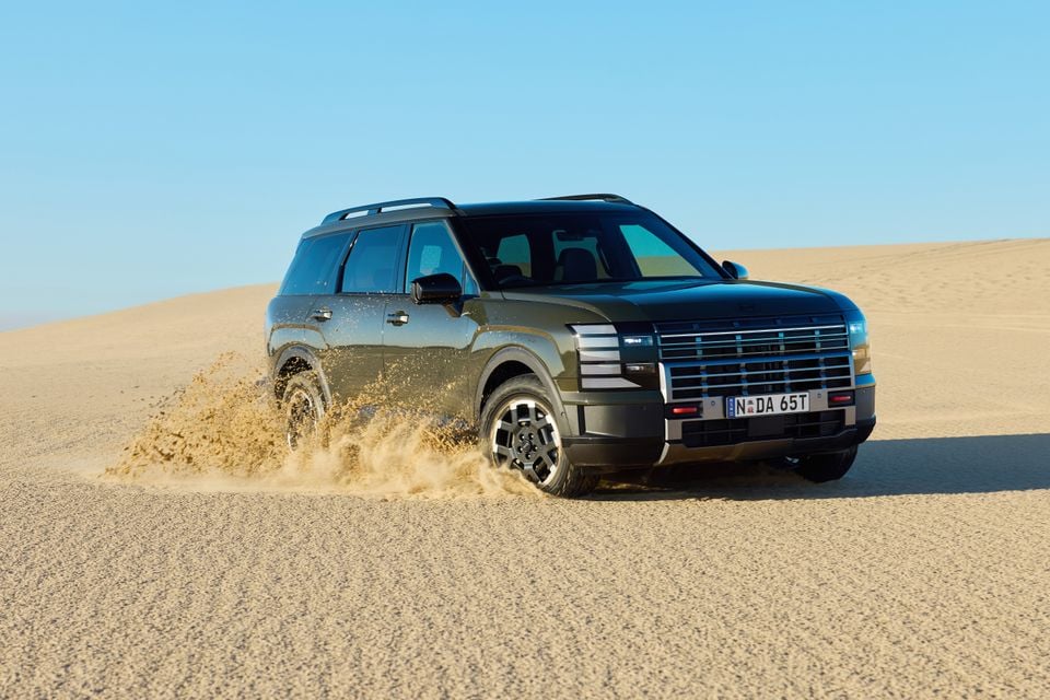 A dark green 2026 Hyundai Palisade XRT Pro large SUV driving through sand dunes, seen from the front-side with sand spraying from the wheels