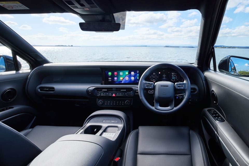 Interior of a Hyundai Palisade showing the dashboard, wide infotainment touchscreen with Apple CarPlay, flat-bottom steering wheel, and dark upholstery, with a coastal water view through the windscreen.