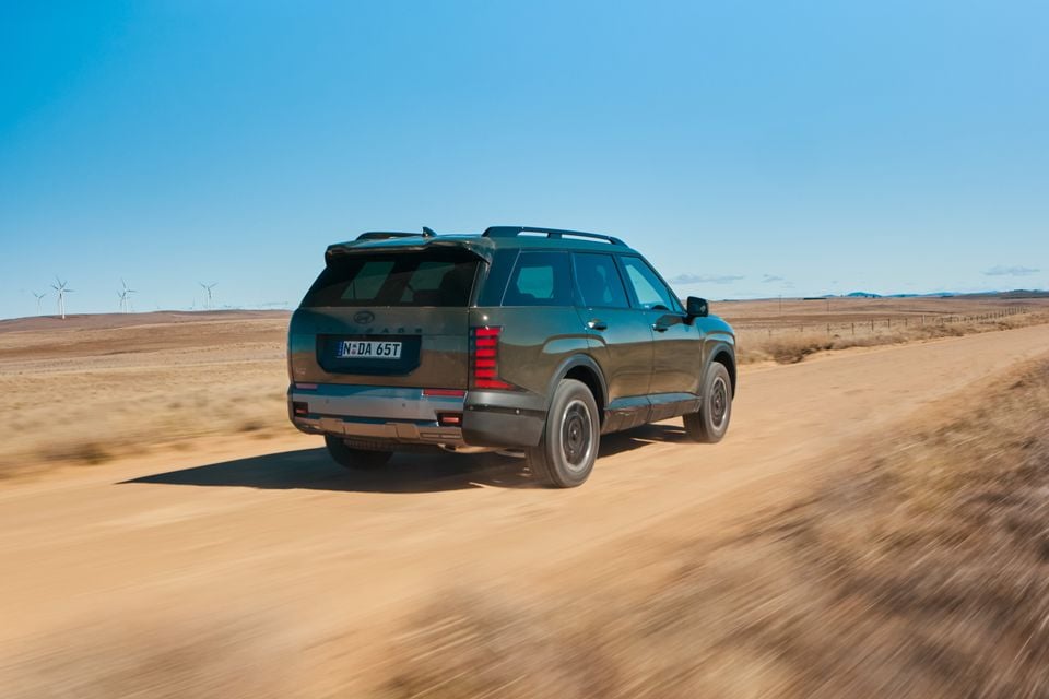 A dark green 2026 Hyundai Palisade XRT Pro SUV driving along a dirt road in an arid Australian landscape, seen from the rear three-quarter angle.