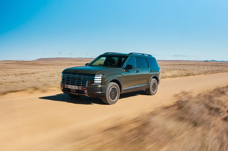 A dark green 2026 Hyundai Palisade XRT Pro large SUV driving along a dusty outback road with wind turbines in the background