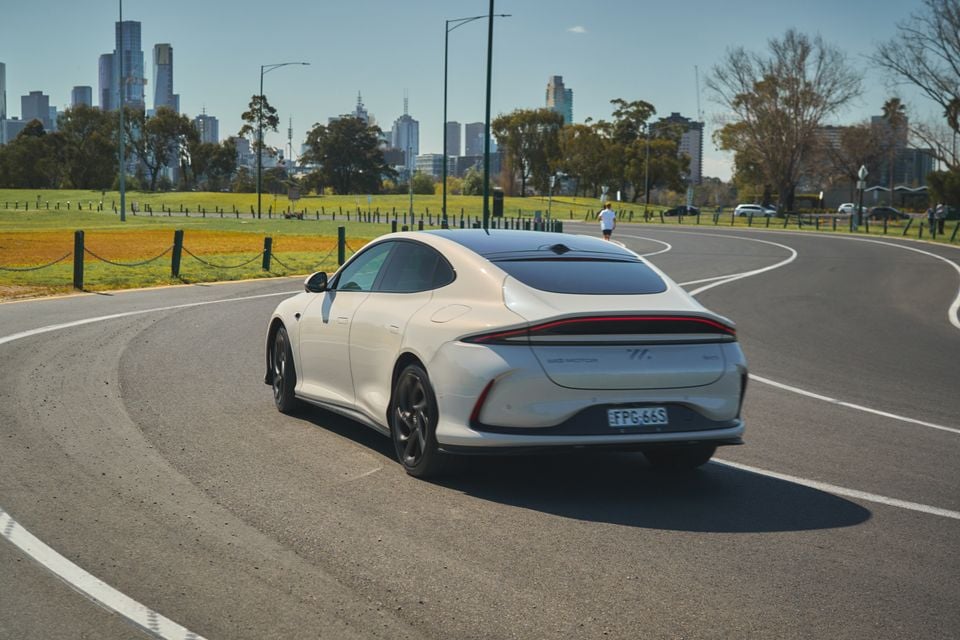 Um sedã IM5 2026 branco dirigindo em uma estrada curva, visto de trás, com o horizonte de uma cidade e um parque atrás