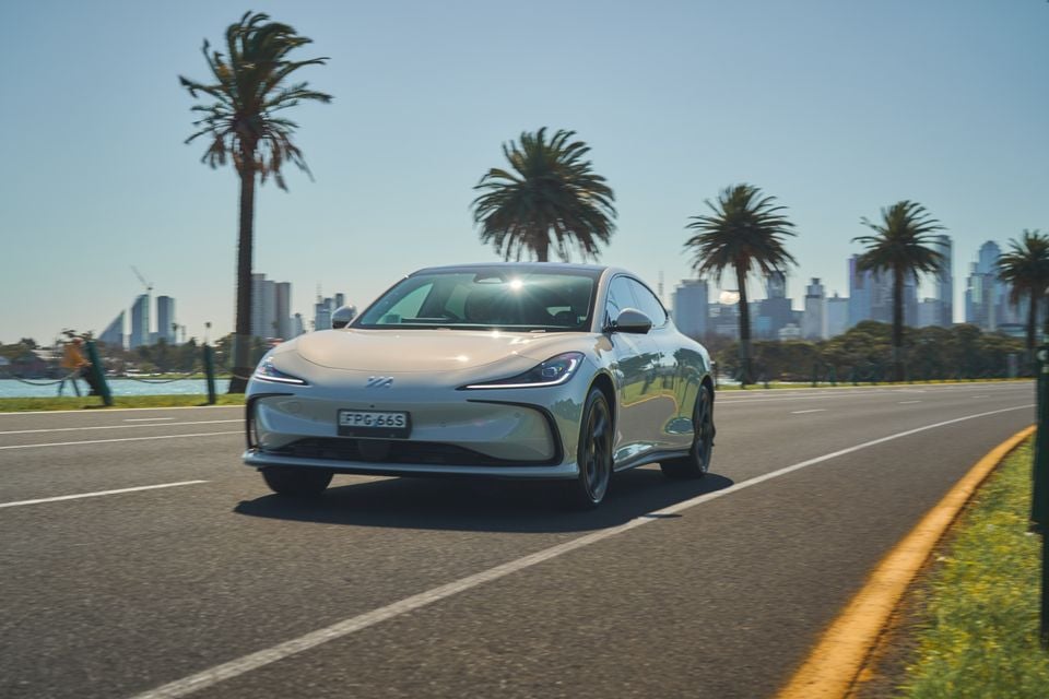 A white 2026 IM5 electric sedan driving along a palm-lined waterfront road with a city skyline behind, seen from the front.