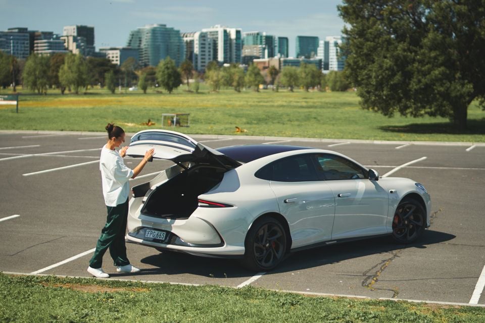 Um liftback IM5 2026 branco visto da parte traseira enquanto uma mulher abre a porta traseira em um estacionamento ao lado do parque com torres urbanas atrás