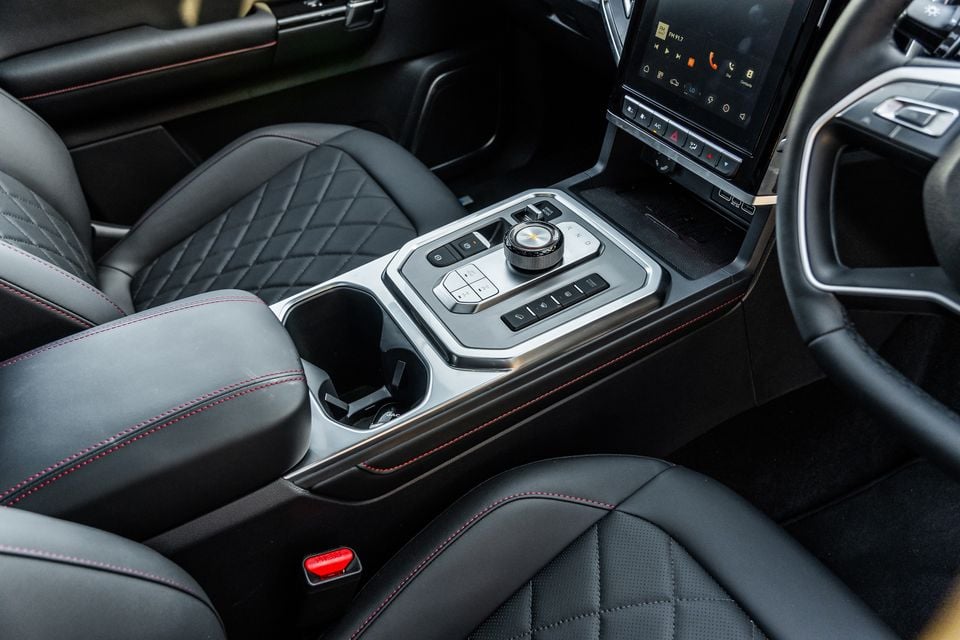 Interior centre console of the 2026 JAC Hunter PHEV, showing a rotary gear selector, control buttons, cup holders, and a touchscreen infotainment display, with quilted black leather seats featuring red stitching