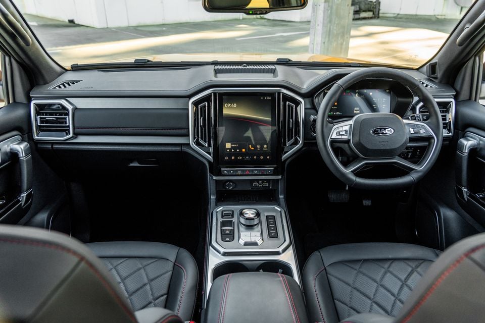 Interior of the JAC Hunter PHEV X showing the dashboard, large central touchscreen infotainment system, steering wheel, digital instrument cluster, and quilted leather seats with red stitching