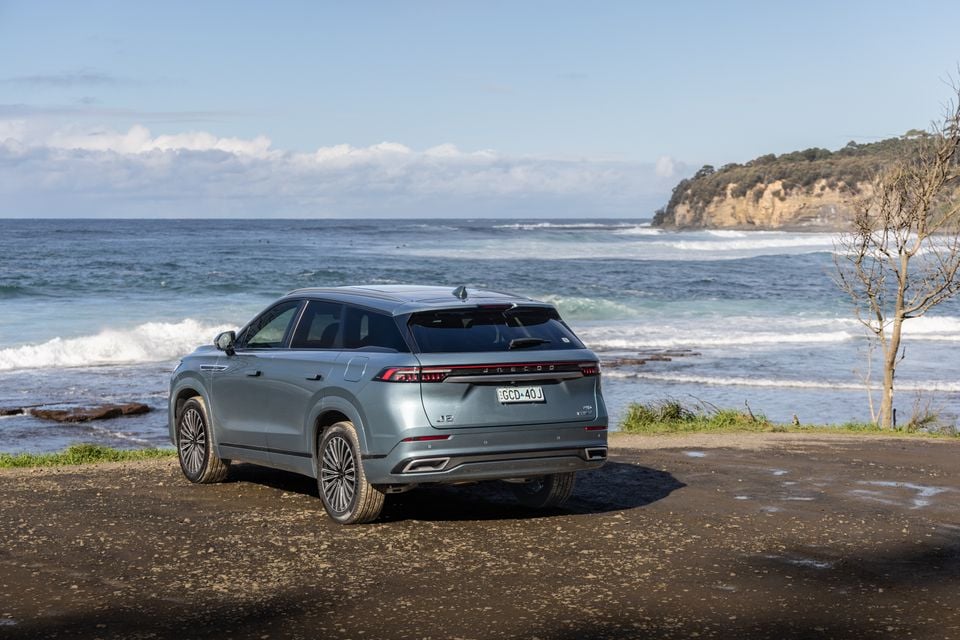 A silver-blue Jaecoo J8 SUV seen from the rear three-quarter angle, parked near a rocky coastal shoreline with ocean waves in the background.