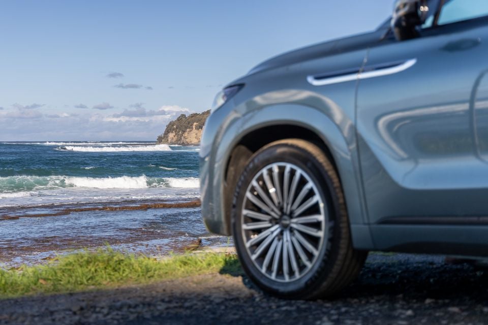 A silver Jaecoo J8 SUV parked near a rocky coastline with ocean waves in the background, seen from a low front-side angle highlighting the front wheel and bumper.