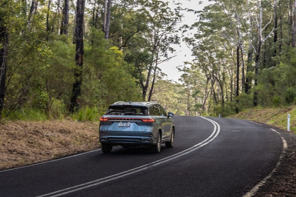 A blue-grey Jaecoo J8 SUV driving away on a winding road through an Australian bush setting, seen from the rear