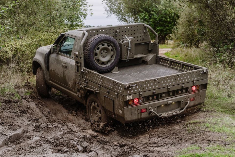 A military-green Land Rover Defender Wolf Series II utility pickup drives through deep mud on a forest trail, seen from the rear quarter.