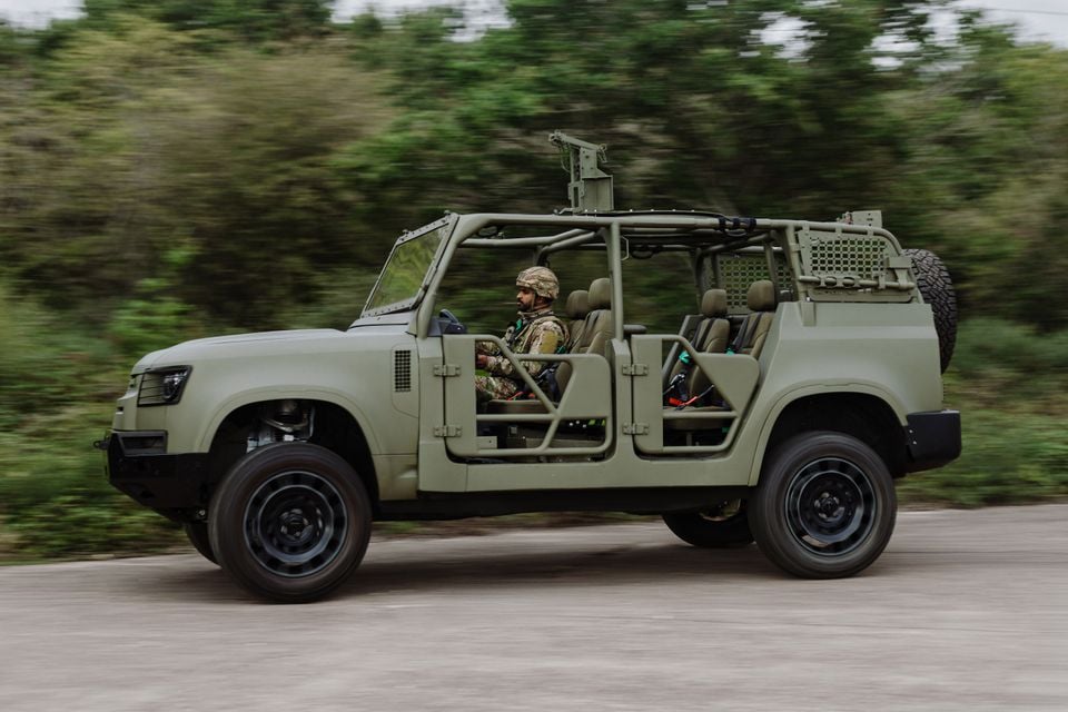 A military-green Land Rover Defender Wolf trials utility vehicle, doors removed and roll cage exposed, driven by a soldier along a road with a tyre mounted on the rear.
