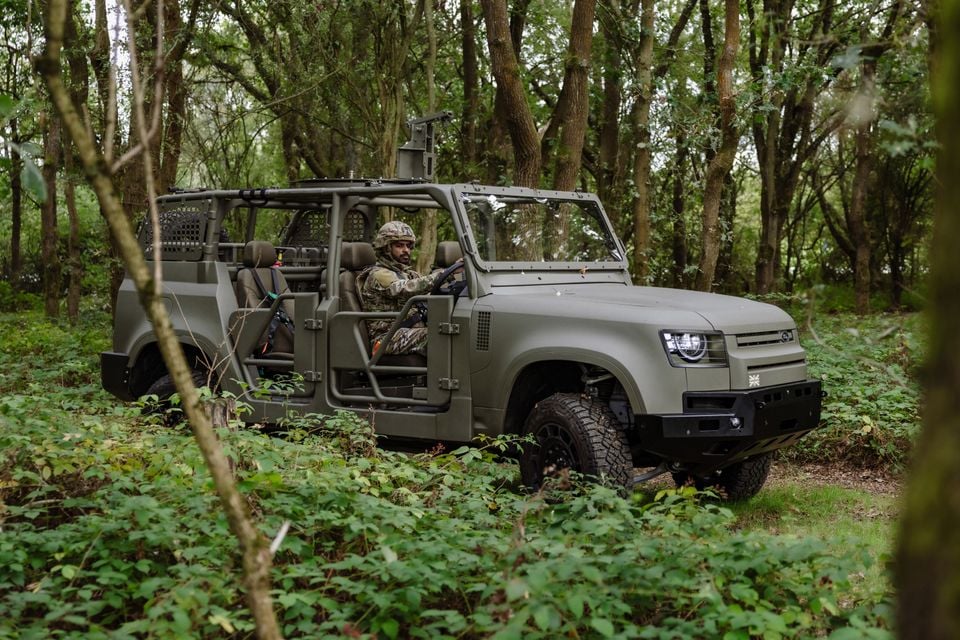 A military-spec Land Rover Defender Wolf, doorless with a roll cage, driven by a soldier through a forest trail.