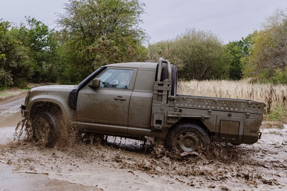 A military-green Land Rover Defender Wolf trials utility pickup truck splashing through a muddy off-road track, seen from the side.