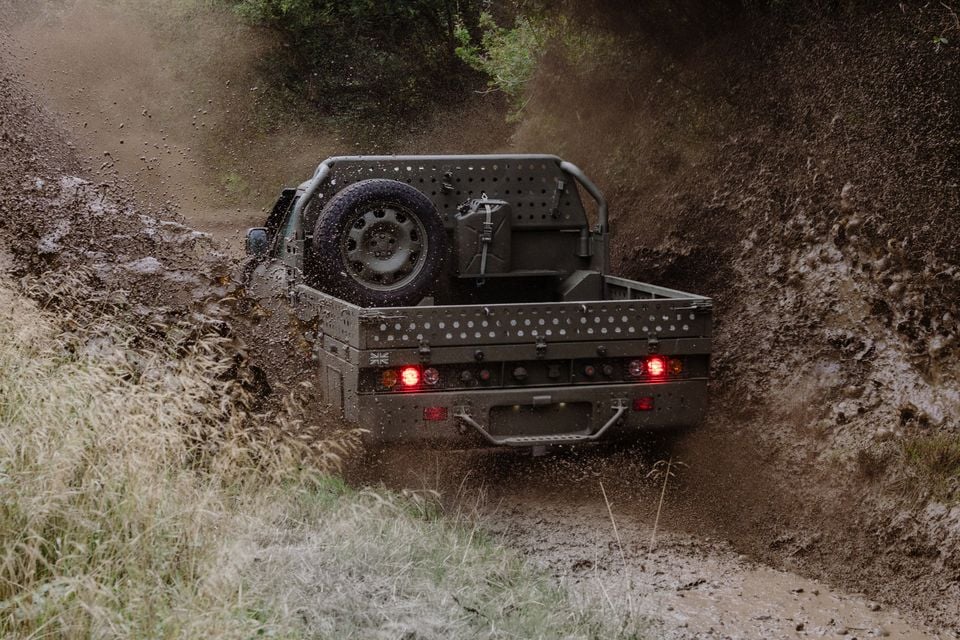 A military-style Land Rover Defender Wolf utility truck splashes through a muddy trail, seen from the rear with its spare wheel mounted in the tray.