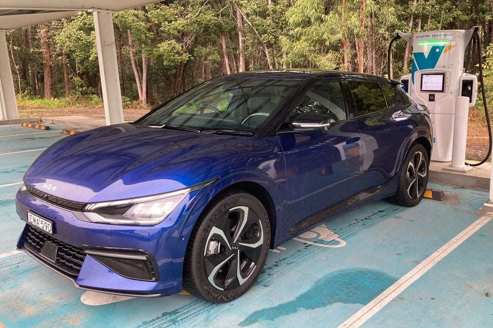 A blue Kia EV6 parked at a public EV charging station, seen from the front-side angle, surrounded by bushland