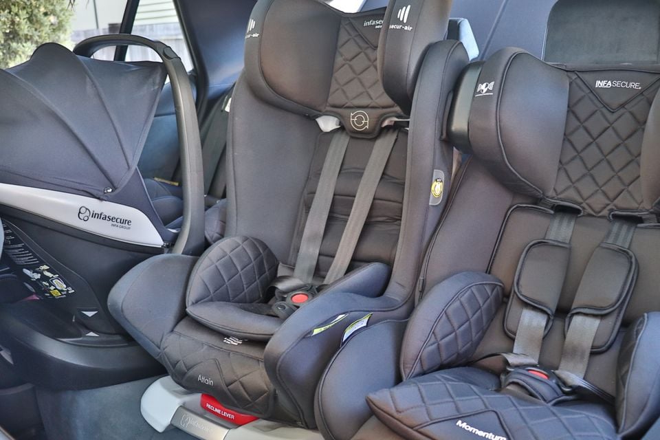 Three child car seats fitted side by side across the rear seat of a car, including Infasecure infant carrier, Attain convertible seat, and Momentum booster seat