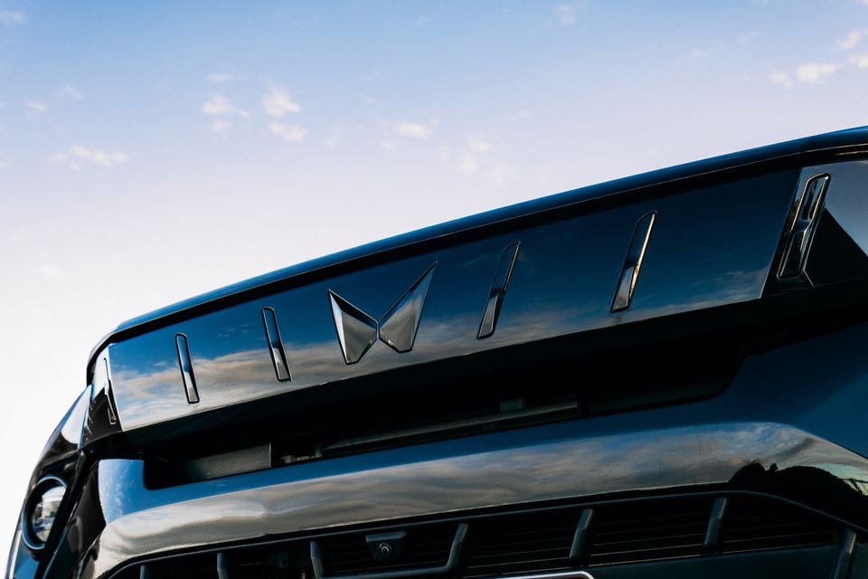 Close-up of the front grille and badge area of a 2026 Mahindra XUV3XO Black Edition, showing the dark emblem and vented panel against a blue sky