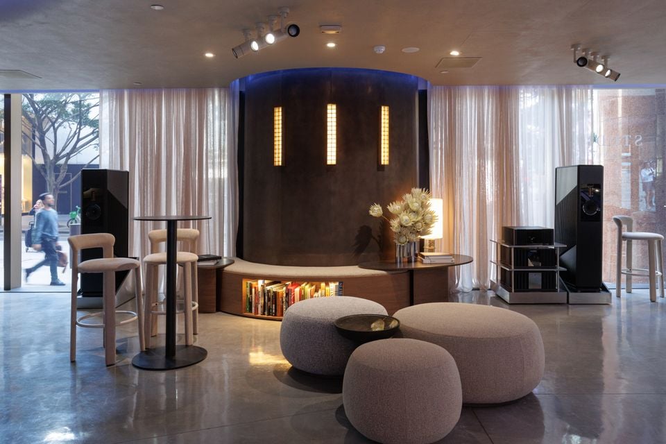 The interior of the Mercedes-Benz studio showroom in Sydney, featuring a curved feature wall with warm lighting, ottomans, bar stools, books, and floor-standing speakers on a polished concrete floor.