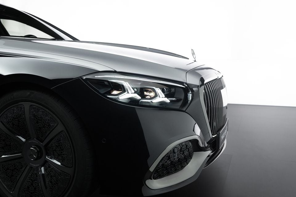 Close-up of the front corner of a black Mercedes-Maybach showing the LED headlight, vertical chrome grille, Maybach badge, and distinctive alloy wheel design against a white studio background.