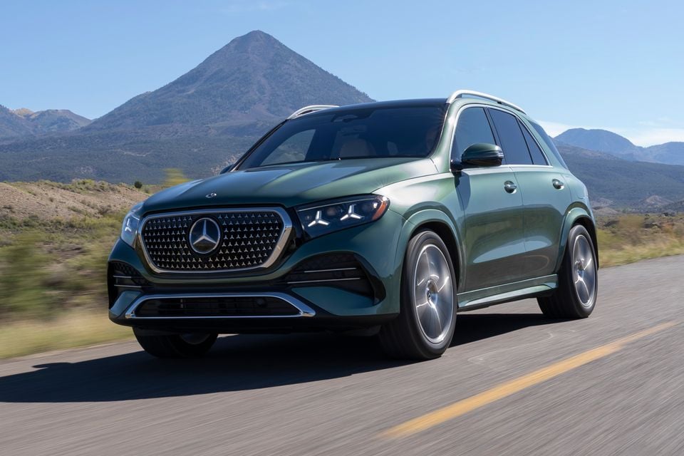 A green 2026 Mercedes-Benz GLE 450e plug-in hybrid SUV driving on a country road with mountains behind, seen from the front