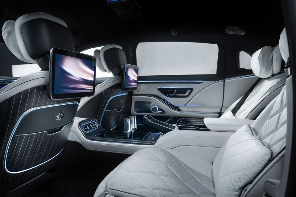 Luxurious rear cabin of a Mercedes-Maybach showing white quilted leather seats, rear entertainment screens, ambient blue lighting, and champagne flutes in the centre console