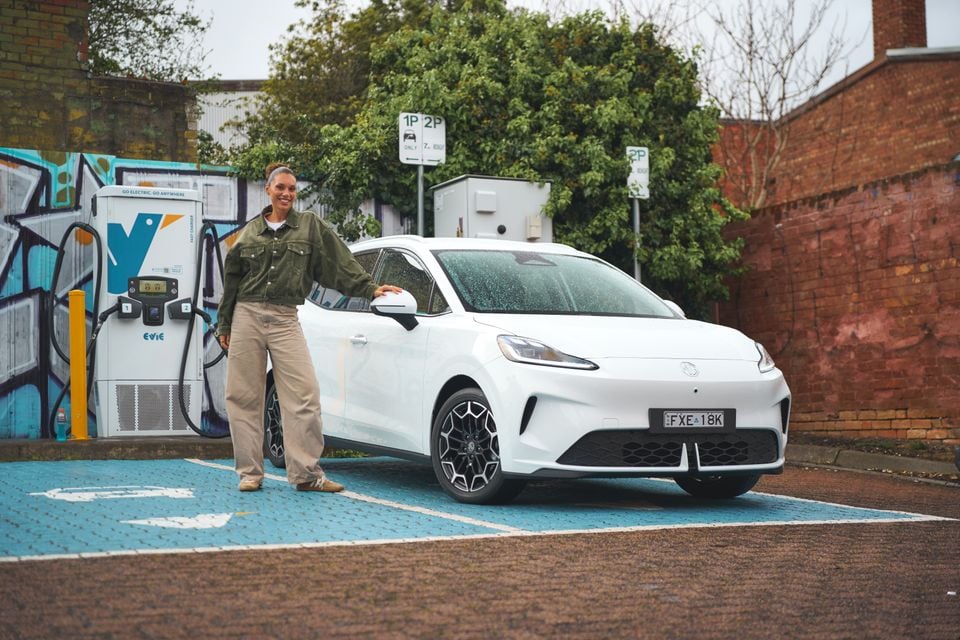 A woman stands next to a white 2026 MG4 EV hatchback parked beside a public EV fast charger.