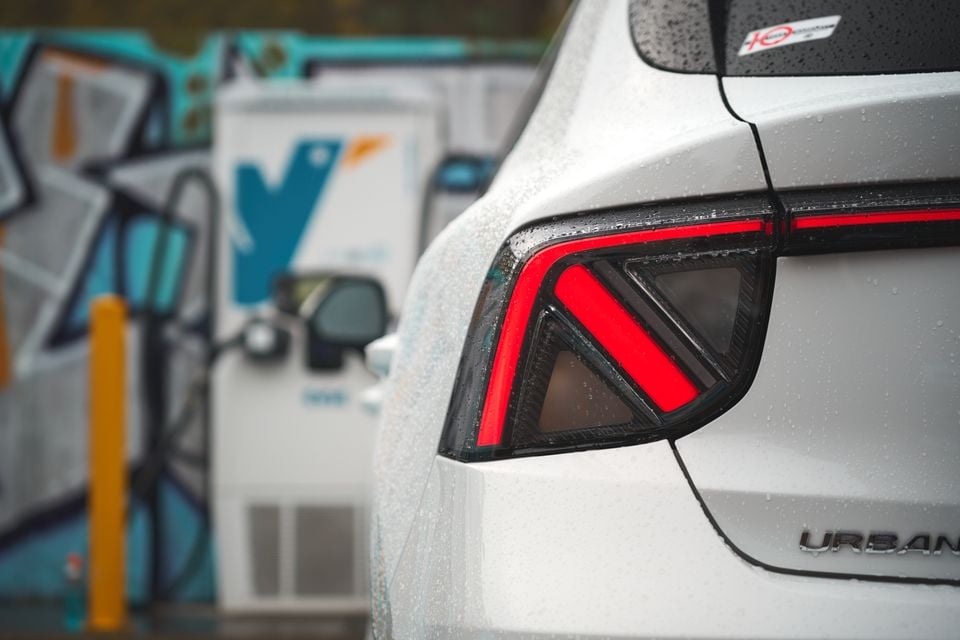 Close-up of the rear taillight on a white 2026 MG4 EV Urban hatchback, with an EV charging station in the background.