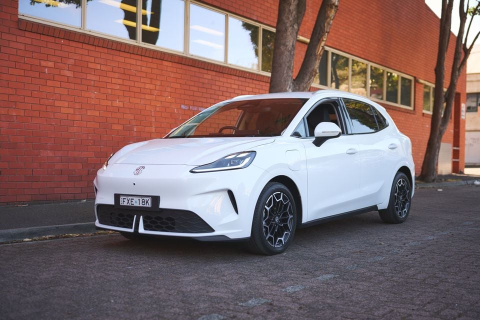A white 2026 MG4 EV hatchback parked on a paved area in front of a red brick building, seen from the front quarter.
