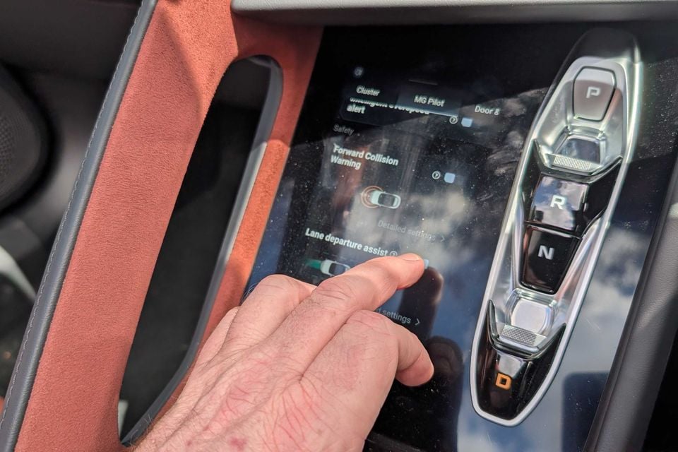 A hand touching the 2026 MG Cyberster's centre touchscreen showing driver assist settings, beside the gear selector and orange suede trim