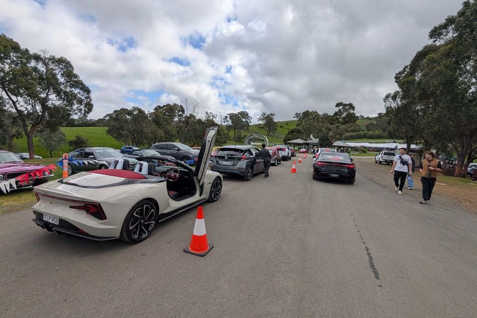 A white 2026 MG Cyberster convertible with its scissor door raised, seen from the rear, queued with other cars at the Rob Roy Hillclimb event