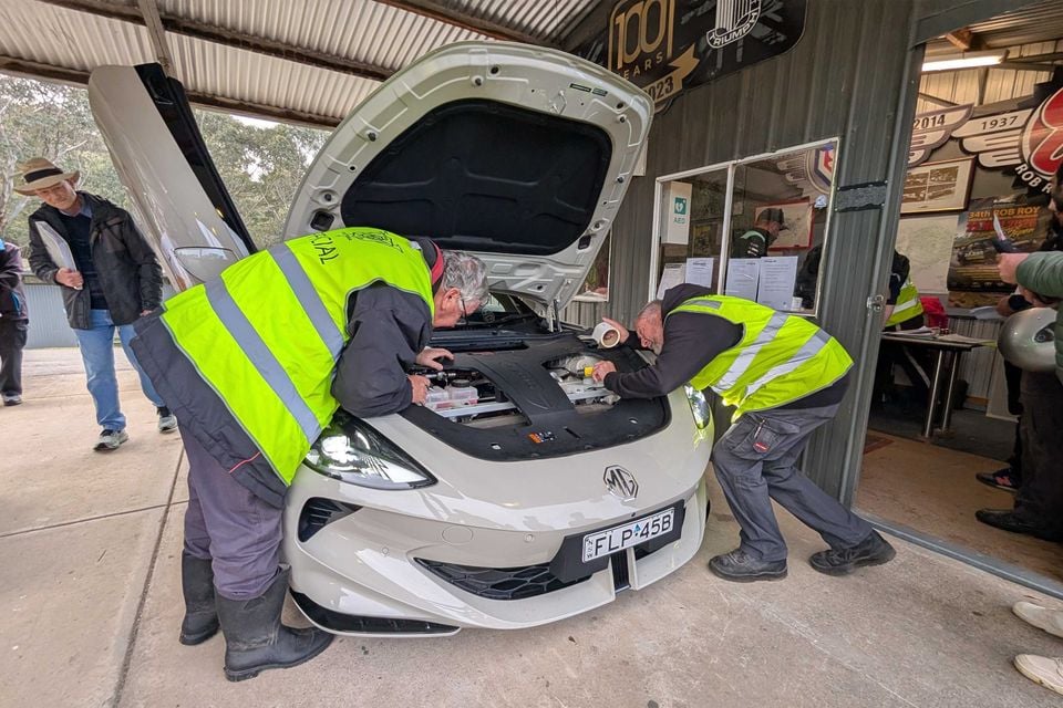 Scrutineers in hi-vis vests inspect under the open bonnet of a white 2026 MG Cyberster at the Rob Roy Hillclimb, with its scissor door raised