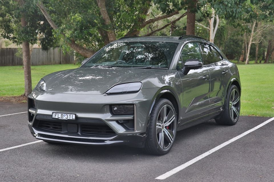 A grey 2026 Porsche Macan SUV seen from the front-side angle, parked in a car park with trees and grass in the background.