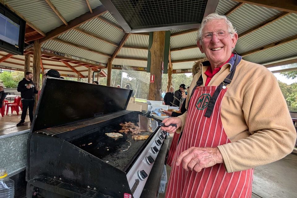 A smiling older man in a red striped MG apron cooks bacon on a barbecue hotplate under a shelter at the Rob Roy Hillclimb