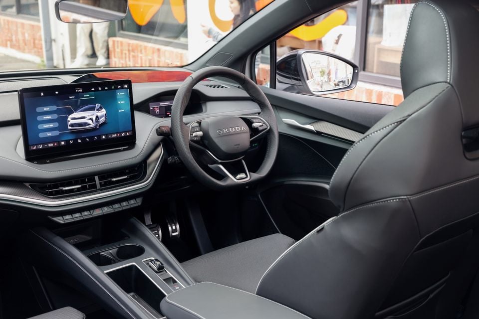 Interior of a Skoda Enyaq showing the steering wheel, large central touchscreen infotainment display, and grey leather seats