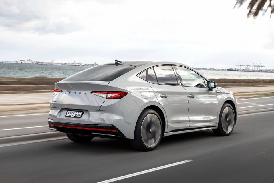 A silver Skoda Enyaq liftback seen from the rear three-quarter angle while driving along a coastal road