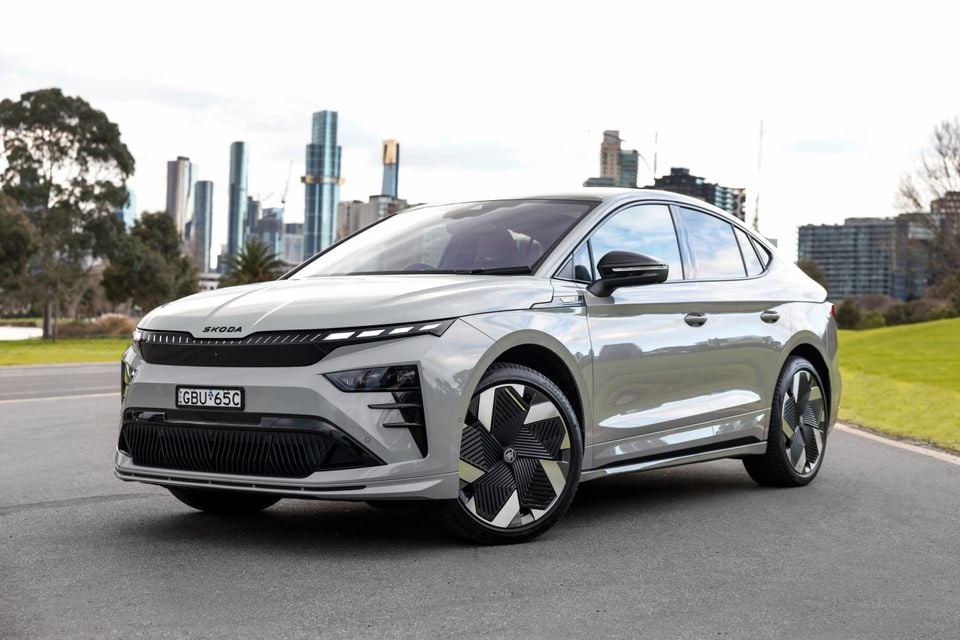A silver 2026 Skoda Enyaq fastback SUV seen from the front three-quarter angle, parked on a road with the Melbourne city skyline in the background.