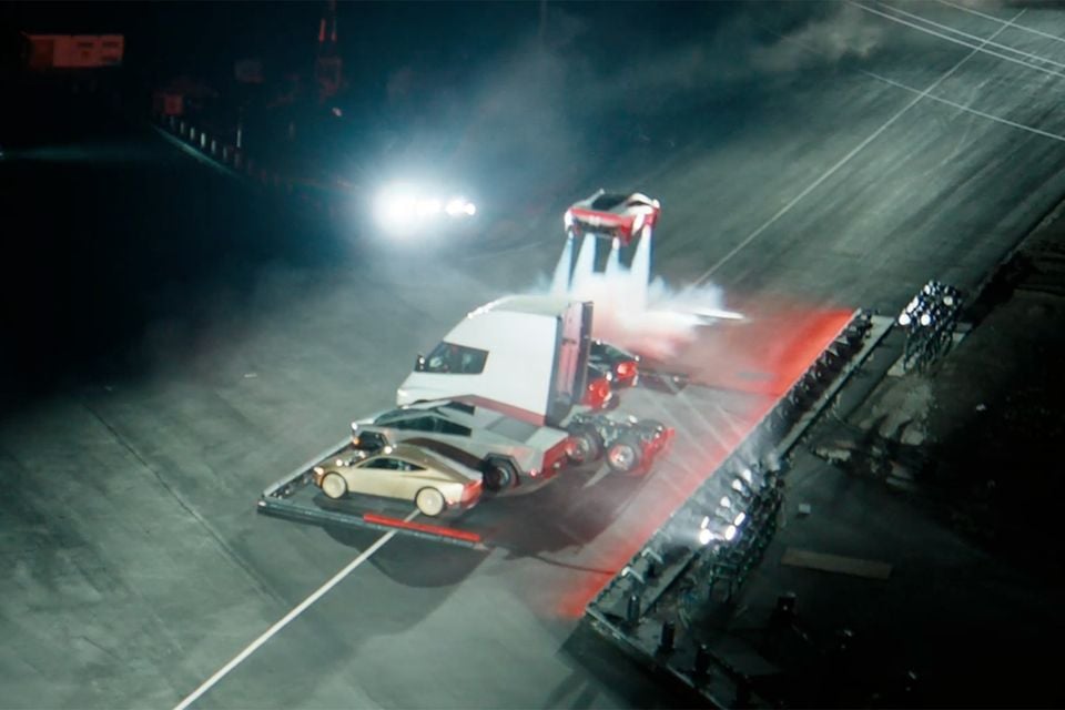 A red Tesla Roadster hovering on thrust jets above a lineup of a Tesla Semi, Cybertruck and gold Cybercab on a dark track at night