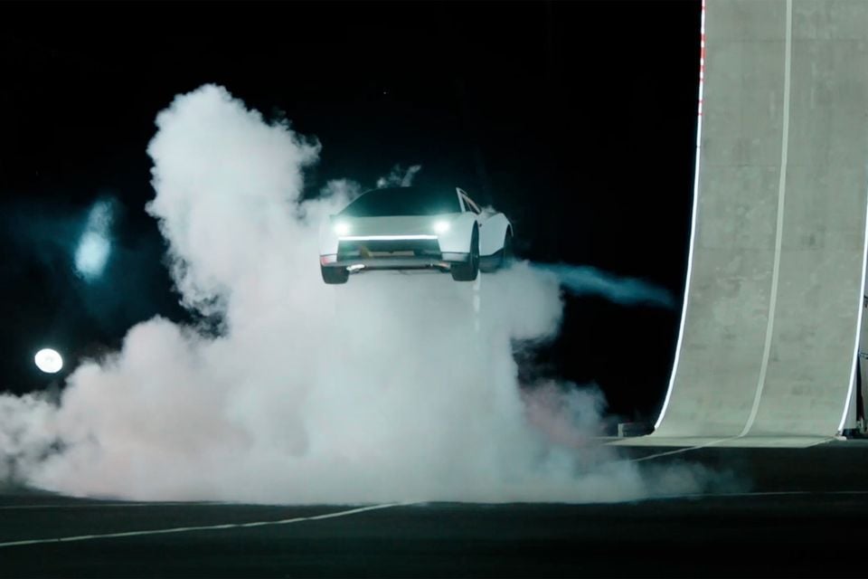 A white Tesla Roadster airborne in a cloud of smoke beside a tall curved ramp at night, seen from the front